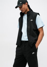 Fekete bolyhos mellény cipzáros zsebbe, világoskék ing fölött viselve. Fekete bolyhos nadrág és sapka, mindkettőn a fehér Nike logo látható.