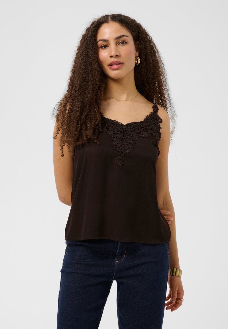 Femme aux longs cheveux bouclés portant un haut sans manches noir orné de dentelle et un jean bleu foncé sur un fond uni.