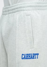Ανοιχτό γκρι παντελόνι φόρμας από μαλακό ύφασμα. Διαθέτει ελαστική μέση, πλαϊνή τσέπη και έντονο μπλε λογότυπο "CARHARTT" στ ο πόδι.
