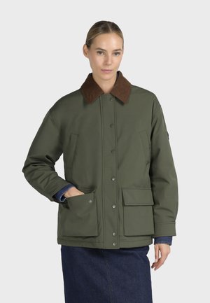 Femme portant une veste vert olive avec un col marron et de grandes poches avant, avec une jupe en denim sur un fond clair uni.