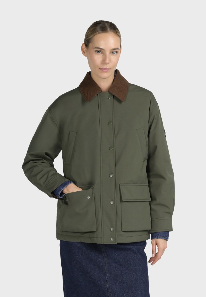 Femme portant une veste vert olive avec un col marron et de grandes poches avant, avec une jupe en denim sur un fond clair uni.