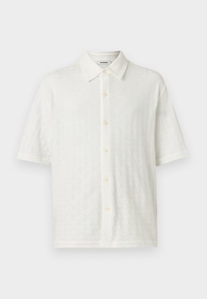 Junger Mann steht mit den Händen in den Taschen, trägt ein strukturiertes weißes Kurzarmhemd, schwarze weite Hosen und beige Loafers.