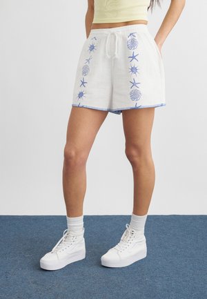 Isik, kes kannab valgeid tavalise paelaga lühikesi pükse siniste mereloomade mustriga, valgeid sokke ja valgeid platvorm-tenniseid, seisab sinisel vaibal.