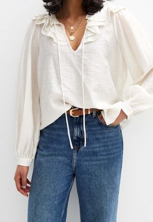 Nainen pukeutunut valkoiseen röyhelöiseen puseroon, jossa on pitkät hihat, paita on niiden yläpuolelle tungettu korkeavyötäröisiin sinisiin farkkuihin, asusteina kerrokselliset kultakaulakorut ja ruskea vyö.