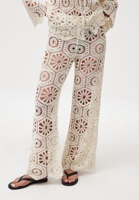 Pantaloni all'uncinetto color crema con motivo floreale geometrico, design a gamba larga e trama a rete. Abbinati a ciabatte nere.