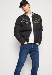 Svart puffjacka med USA- och bokstav B-broderi, ribbat krage med vita och orange detaljer, bärs med blå jeans.