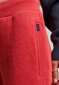 Red textured sweatpants pocket with small navy blue Superdry label, next to a hand wearing gold rings and a navy blue sleeve.