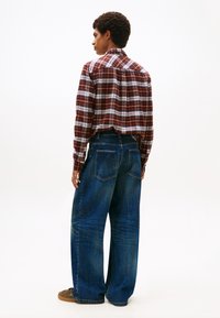 Camicia a quadri rossa, bianca e nera con maniche lunghe; abbinata a jeans larghi di colore blu scuro con grandi tasche posteriori.