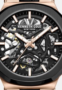 Kenneth Cole automatische horloge met een zwarte skeleton wijzerplaat, rosegouden accenten, zwarte bezel en een gestructureerde zwarte band.