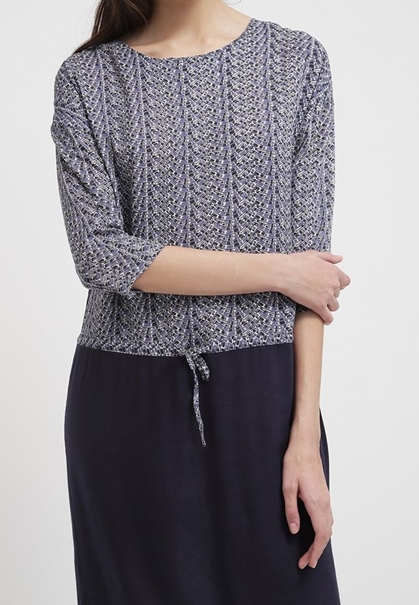 Femme portant une robe avec un haut gris et blanc à motifs avec des manches 3/4 et une jupe unie bleu marine foncé nouée à la taille.