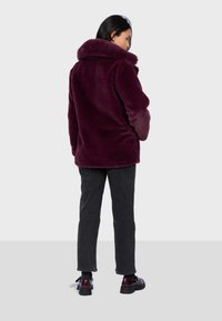 Mujer con chaqueta de piel sintética púrpura oscuro, pantalones negros y zapatos burdeos oscuros, de pie de espaldas frente a un fondo gris claro liso.