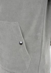 Grijze corduroy stof met een gestructureerd oppervlak, voorzien van een schuin zakje en een zilveren drukknoopdetail voor extra functionaliteit.