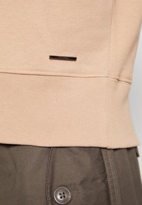 Dobladillo de sudadera beige con una placa de logo metálica, combinado con pantalones marrón oscuro que presentan un botón visible y detalles de costura.