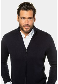 Mörkblå zippered cardigan tillverkad av texturerat stickat tyg, med hög krage och ribbade ärmmuddar. Stylad över en vit skjorta med krage.