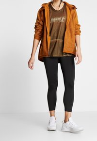 Sudadera con cremallera de forro polar naranja, camiseta gráfica marrón, leggings negros y zapatillas deportivas blancas llevadas por una figura de pie.