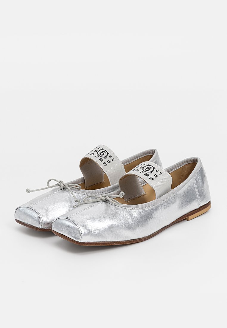 Ballerine metalliche argento con punte quadrate, piccoli fiocchi sulla parte superiore e cinturini elastici bianchi con numeri neri e un "6" cerchiato.