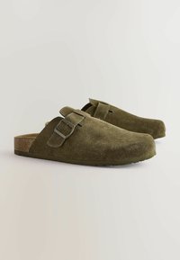 Olive green suede clogs with a rounded toe, adjustable buckle strap, and cork sole. Textured surface, minimalist design, and slip-on style.