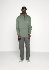 Maslinasto zelena majica s kapuljačom i prednjim džepom te crnom vrpcom za vezanje, uparena s sivim cargo hlačama i zelenim kliznim cipelama s karo uzorkom.