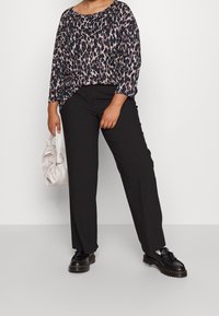 Leopardenmuster-Oberteil in dunklen Tönen, kombiniert mit schwarzen Hosen. Das Model hält eine hellbeige Tasche und trägt schwarze Schuhe mit einer dicken Sohle.