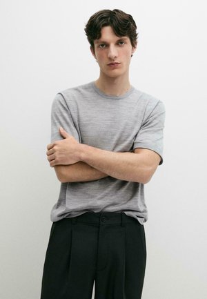 Giovane uomo con capelli scuri che indossa una camicia grigio chiaro a maniche corte e pantaloni neri, in piedi con le braccia incrociate su sfondo bianco.