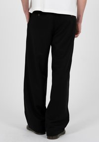 Person trägt schwarze, weit geschnittene Hose und schwarze Schuhe, die Hände ruhen in den hinteren Taschen, steht vor einem schlichten, hellen Hintergrund.