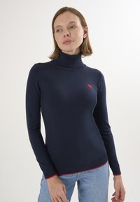 Maglione a collo alto navy con un emblema a forma di cuore rosso sul petto e dettagli rossi sull'orlo. Realizzato in materiale morbido e texture. Design aderente.