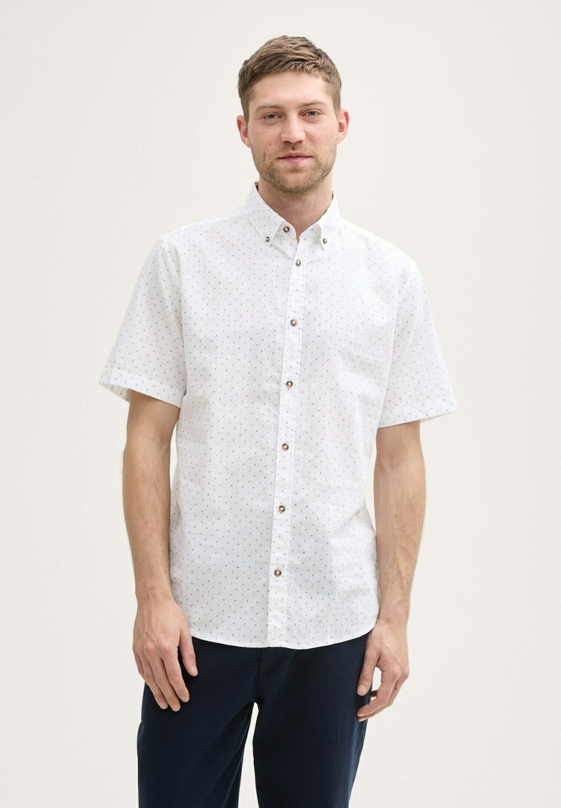 Chemise blanche à manches courtes avec des motifs de pois colorés, col boutonné, et boutons marron, fabriquée en tissu léger. Portée avec un pantalon marine.