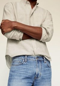 Chemise en lin gris clair avec col boutonné, manches longues retroussées jusqu'aux coudes, associée à un jean en denim bleu délavé présentant un effet whiskering.