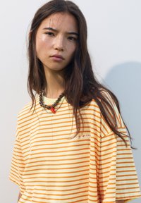 T-shirt rayé avec des lignes jaunes et oranges, présentant une coupe décontractée et des manches courtes. Comprend un petit détail brodé. Collier circulaire.
