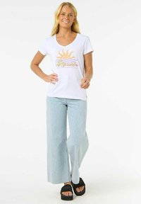 Hvid kortærmet t-shirt med et solgrafik i gul og orange, parret med lyseblå brede jeans og sorte platform-sandaler.