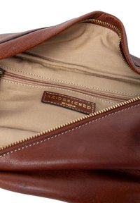 Brun lædertaske indvendig med beige foring, der har en lynlåslomme og et synligt mærke med teksten "REDESIGNED."