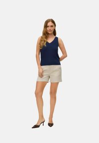 Top in maglia a coste di colore blu navy con scollatura a V, abbinato a pantaloni beige. L'aspetto è completato da mule con tacco marroni con dettagli decorativi.