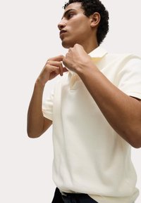 Cremefarvet ribbet polo t-shirt med korte ærmer, tre-knap lukning og en blød tekstur, parret med mørke bukser.