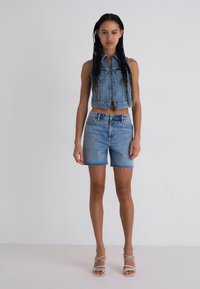 Gilet in denim e pantaloni corti a vita alta in blu chiaro, con orli sfrangiati. Modella in piedi a piedi nudi, che mostra sandali con lacci.