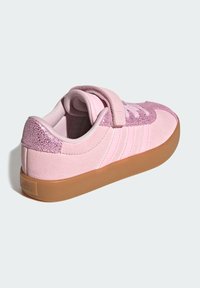 Pink suede shoe with a textured pink accent, featuring a velcro strap, rounded toe, and a brown rubber sole. Three stripes on side.