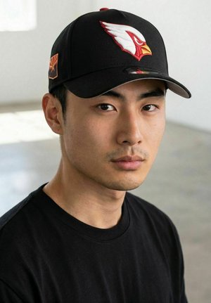 Junger Mann mit schwarzer Kappe, auf der ein rotes und weißes Adler-Logo abgebildet ist, und schwarzem T-Shirt, der mit neutralem Gesichtsausdruck nach vorne schaut.