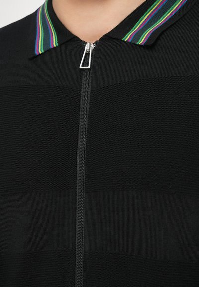 Čierny pletený top na zips s golierom s pruhmi v zelenej, modrej a fialovej farbe. Materiál má textúrovaný povrch s horizontálnymi pruhmi.