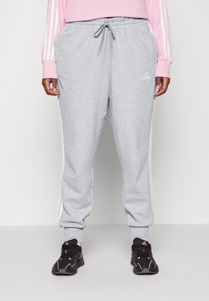 Personne portant un pantalon de survêtement Adidas gris clair avec des bandes blanches, des baskets noires et un haut à manches longues rose, debout devant un fond uni.