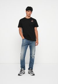 Camiseta negra de manga corta con un gráfico de araña, combinada con jeans azules desgastados y zapatillas en blanco y negro. Fondo liso.