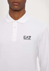 Hvit langermet polo-skjorte laget av bomull, med en svart EA7 Emporio Armani-logo på venstre bryst og knappestolpe med tre knapper.