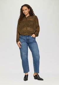 Braun gemusterte transparente Bluse mit Knopfdetails, kombiniert mit blauen gerade geschnittenen Jeans und schwarzen Absatzmules, posierend vor einem hellen Hintergrund.