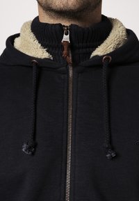 Uomo che indossa una felpa nera con zip, foderata in fleece beige e colletto esterno nero a maglia, con la zip completamente chiusa fino al collo.