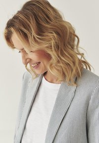 Pruhovaný blazer v modro-bílé látce, spárovaný s jednobarevným bílým tričkem. Modelka má vlnité vlasy po ramena a mírně nakloněnou hlavu.