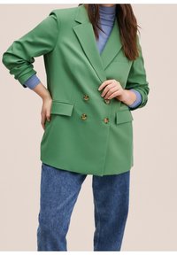 Zielona podwójnie zapięta marynarka w gładkim materiale z poduszkami na ramionach, dużymi klapami i dwiema kieszeniami z przodu. Noszona na jasnoniebieskiej bluzce i dżinsach.
