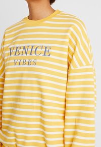 Persona que lleva una camiseta de manga larga a rayas amarillas y blancas con "VENICE VIBES" impreso en el pecho con letras grises.