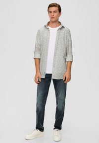 Hellgrau gemusterte Button-Down-Bluse über einem weißen T-Shirt, kombiniert mit dunkelblauen Jeans und weißen Sneakers. Moderner, lässiger Schnitt.