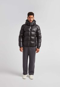 Giacca a vento nera con finitura lucida, cappuccio e zip frontale. Abbinata a pantaloni della tuta grigi e scarpe bianche. Design e texture semplici.