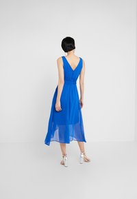 Robe bleue sans manches avec un dos en V, taille cintrée et tissu fluide et transparent atteignant le milieu du mollet. Associée à des sandales à talons argentées.