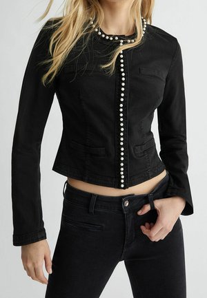 Vrouw draagt een nauwsluitend zwart jasje met parelaccenten langs de voorkant en kraag, gecombineerd met een zwarte broek, tegen een effen achtergrond.