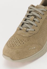 Sneaker in suede color tan con dettagli in rete, punta rotonda e decorazioni forate. Presenta lacci piatti e una suola in gomma per grip. Logo del marchio visibile.